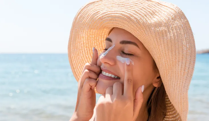 A smiling woman wearing a straw hat applies sunscreen to her face.