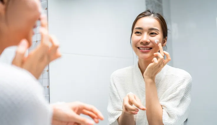 A smiling woman applies a moisturizer to her face.